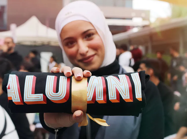 A young woman proudly holding a rolled black fabric tied with a gold ribbon, showcasing the 'ALUMNI' during an AUK event.