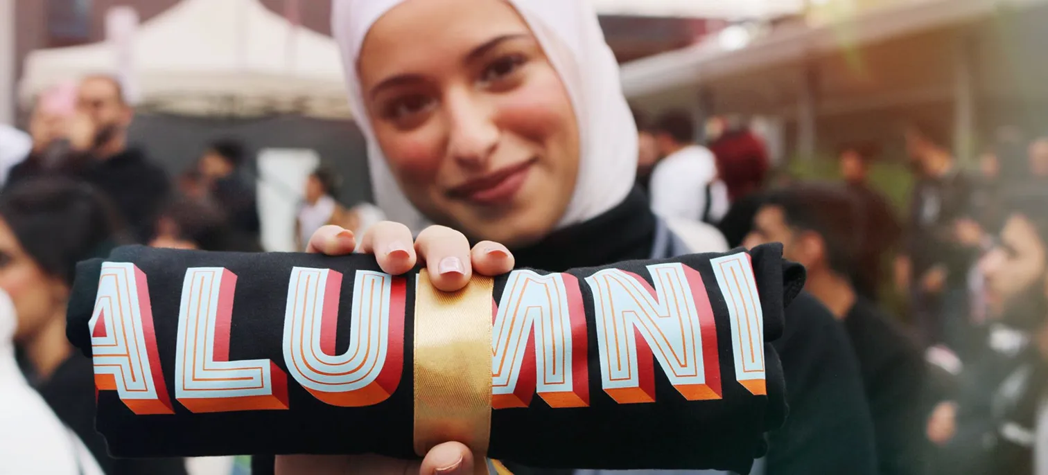 A young woman proudly holding a rolled black fabric tied with a gold ribbon, showcasing the 'ALUMNI' during an AUK event.