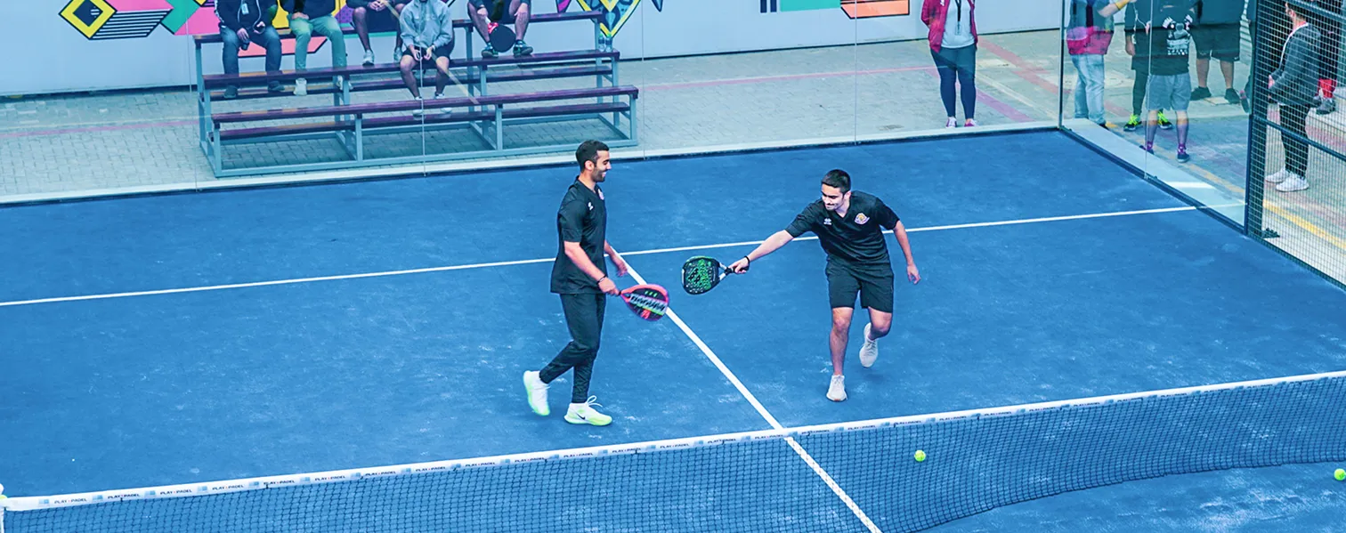 Two AUK students playing doubles tennis, cheered on by a group of their peers on campus.