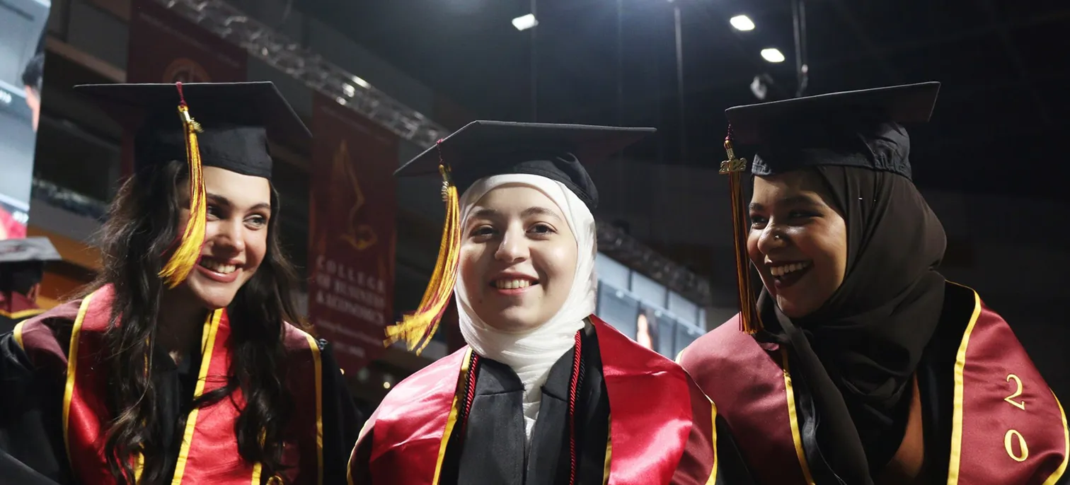 Three AUK graduates joyfully celebrate their achievements as graduates, wearing traditional caps and gowns after their ceremony.