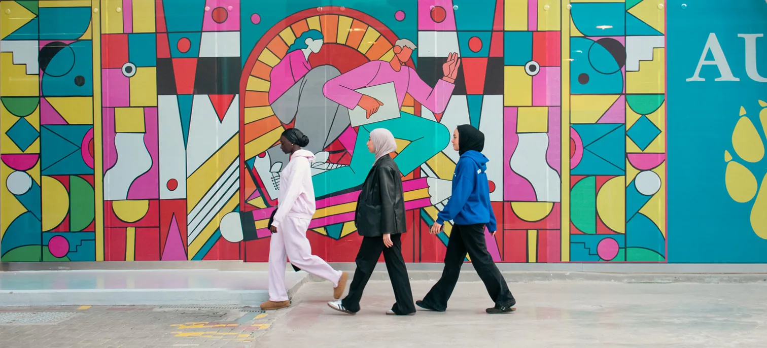 Three AUK students walking on the AUK campus beside wall art.
